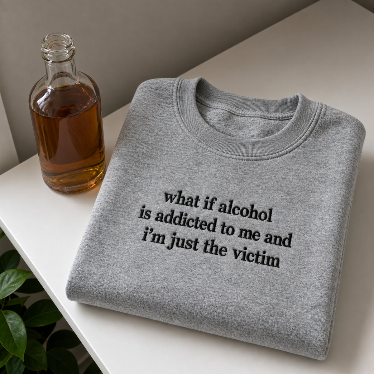 A folded heather gray crewneck sweatshirt with black printed text reading 'what if alcohol is addicted to me and I'm just the victim', styled on a white tabletop next to a glass bottle of amber liquid and a small green plant.