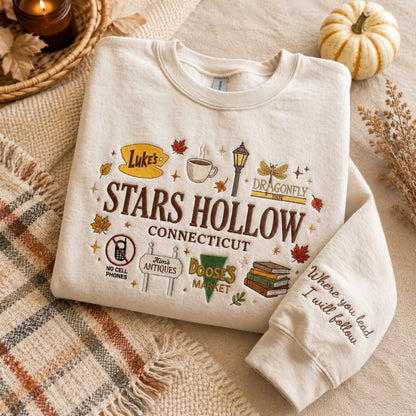 A folded cream sweatshirt with a colorful 'Luke's Stars Hollow Connecticut' graphic, styled on a cozy autumn-themed background with a plaid blanket, dried leaves, a small pumpkin, and a lit candle.