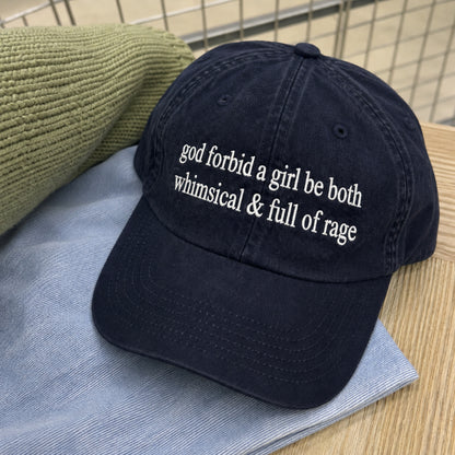 A dark navy blue cotton baseball cap with white embroidered text reading 'god forbid a girl be both whimsical & full of rage', styled on a light blue striped shirt over a green knit fabric on a wooden surface.
