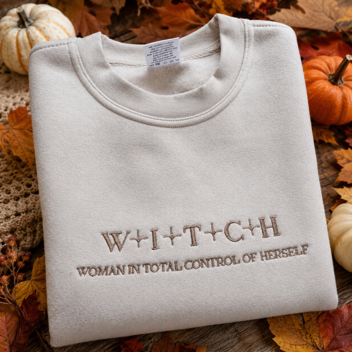 A folded cream-colored crewneck sweatshirt with brown embroidered text reading 'W+I+T+C+H' and 'WOMAN IN TOTAL CONTROL OF HERSELF', styled on a rustic wooden surface with autumn leaves and small pumpkins.