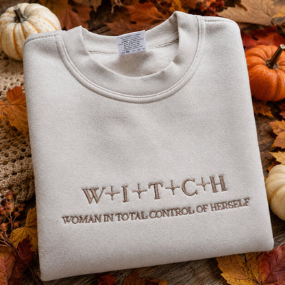 A folded cream-colored crewneck sweatshirt with brown embroidered text reading 'W+I+T+C+H' and 'WOMAN IN TOTAL CONTROL OF HERSELF', styled on a rustic wooden surface with autumn leaves and small pumpkins.