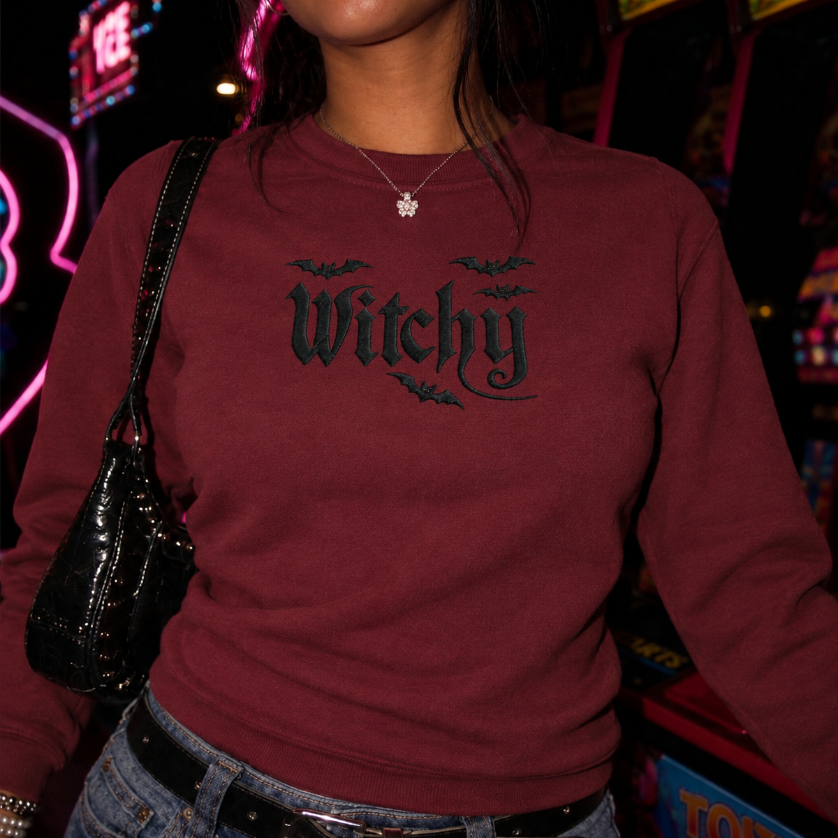 A person wearing a burgundy crewneck sweatshirt with a black gothic-style 'Witchy' graphic and bat motifs on the chest, paired with blue jeans and a black shoulder bag in a neon-lit environment.