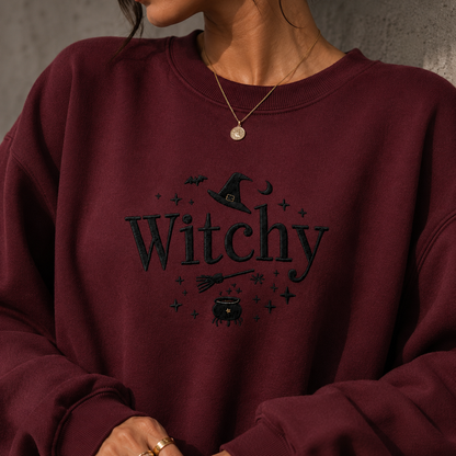 A close-up of a person wearing a burgundy crewneck sweatshirt with a black embroidered graphic that reads 'Witchy' with a witch hat, broom, cauldron, and stars.