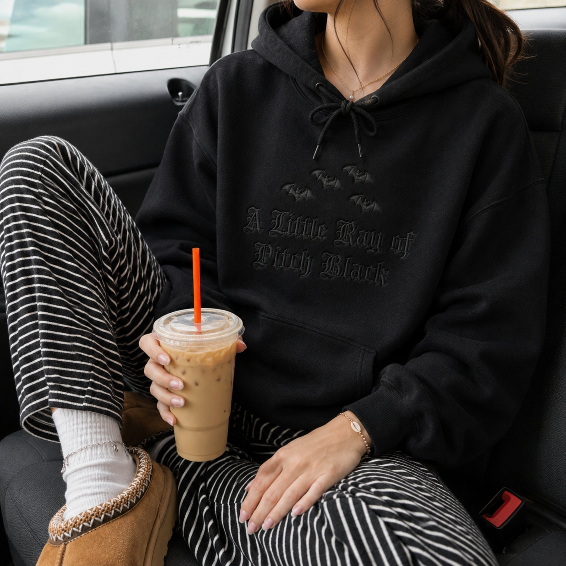 Woman wearing a witchy Halloween crewneck hoodie embroidered with the A Little Ray of Pitch Black gothic design and black bats while relaxing with an iced coffee in casual comfort.