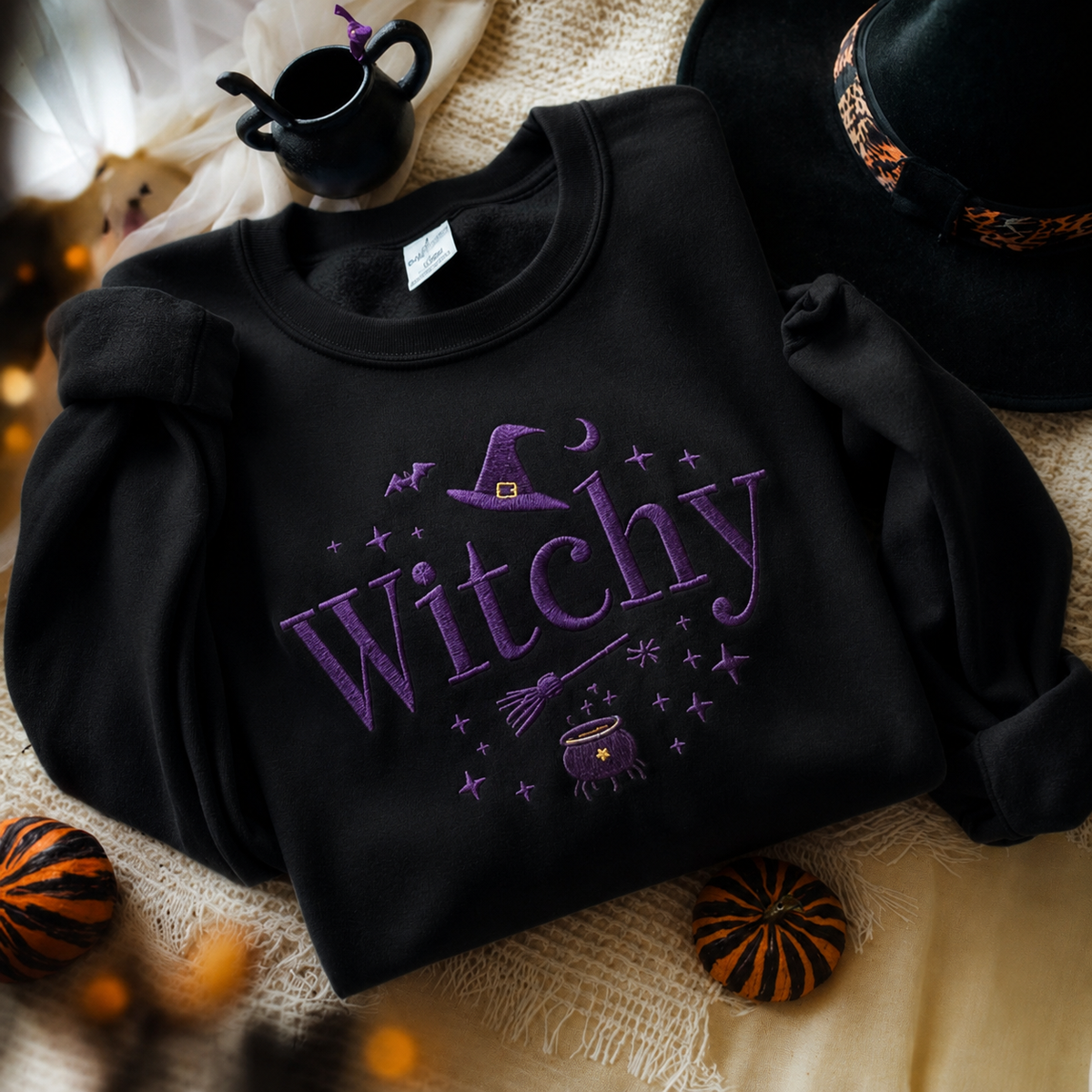 A black long-sleeve sweatshirt with purple embroidered text reading 'Witchy' and a witch hat graphic, styled in a flat lay with a witch hat, small pumpkins, and a black teapot on a beige fabric background.