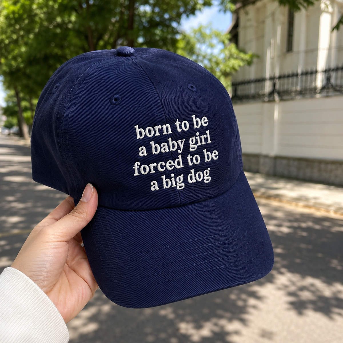 A navy blue baseball cap held by a hand outdoors on a sunny street. The cap features white embroidered text that reads 'born to be a baby girl forced to be a big dog'.