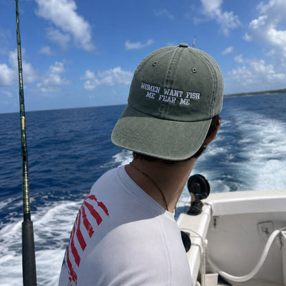 A man on a boat wearing an olive green baseball cap with the embroidered text 'WOMEN WANT FISH ME FEAR ME' in white. The background shows the blue ocean and sky from the moving boat.