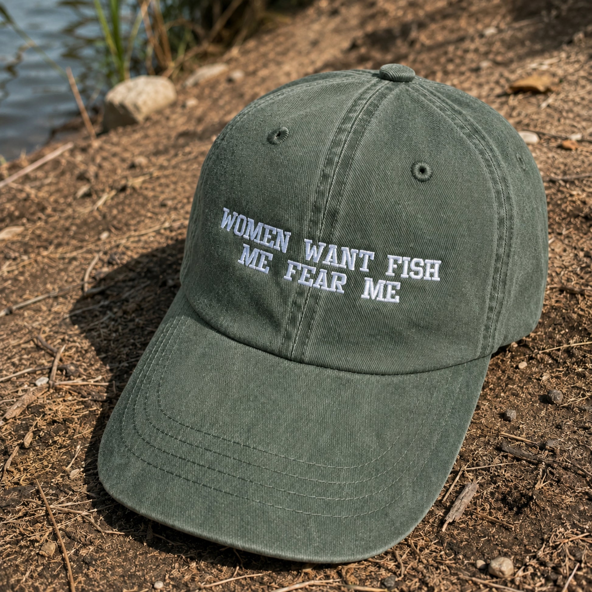 An olive green baseball cap with white embroidered text reading 'WOMEN WANT FISH ME FEAR ME' sits on a dirt bank near water.