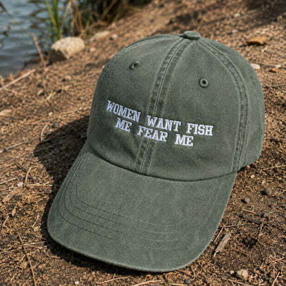 An olive green baseball cap with white embroidered text reading 'WOMEN WANT FISH ME FEAR ME' sits on a dirt bank near water.