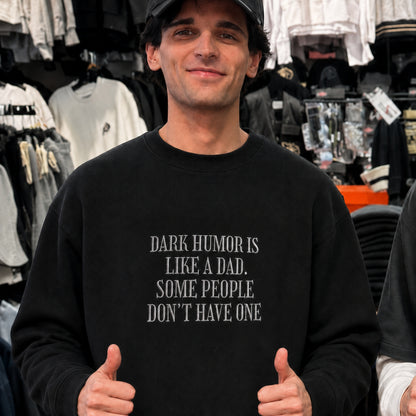 A man wearing a black crewneck sweatshirt with the text 'DARK HUMOR IS LIKE A DAD. SOME PEOPLE DON'T HAVE ONE' in white capital letters, standing in a clothing store.