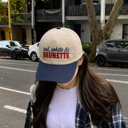 A woman wearing a beige and navy blue baseball cap with 'red, white & BRUNETTE' embroidered on the front, standing on a city street.