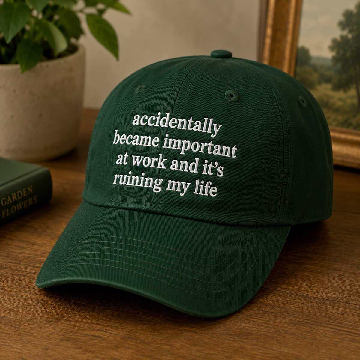 A dark green baseball cap with white embroidered text reading 'accidentally became important at work and it's ruining my life' on a wooden table, with a potted plant and framed picture in the background.