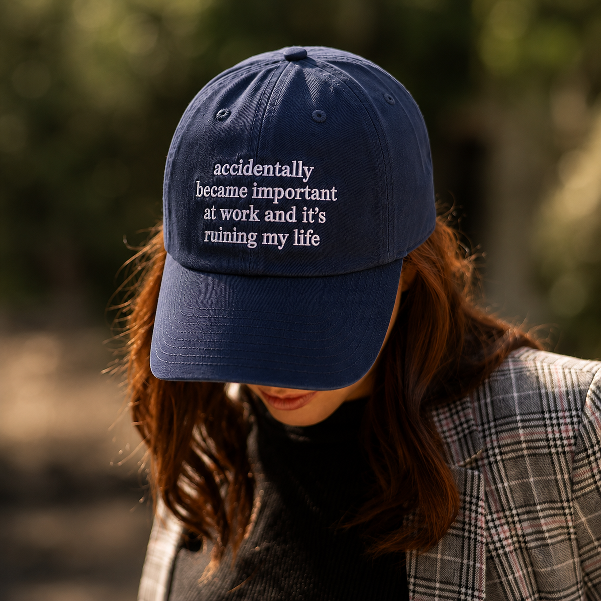 A navy blue baseball cap with white embroidered text reading 'accidentally became important at work and it's ruining my life', worn by a woman in an outdoor setting.