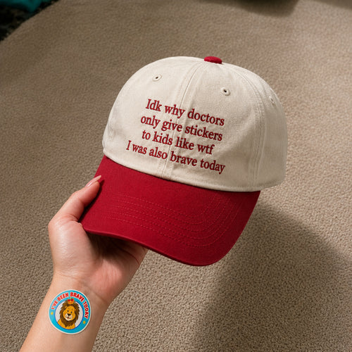 A hand holds a two-tone baseball cap with an off-white crown and a red brim. The cap features red embroidered text that reads, 'Idk why doctors only give stickers to kids like witf I was also brave today.