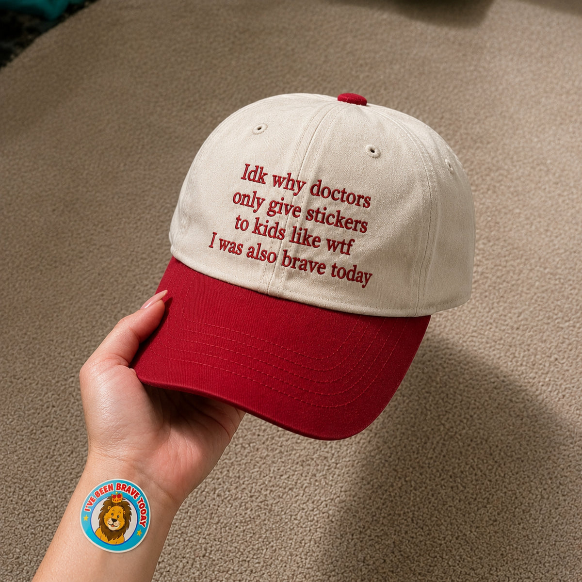 A hand holds a two-tone baseball cap with an off-white crown and a red brim. The cap features red embroidered text that reads, 'Idk why doctors only give stickers to kids like witf I was also brave today.