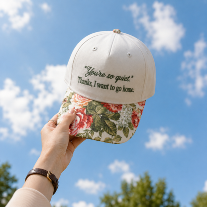 Hand holding a beige baseball cap with black text 'You're so quiet. Thanks, I want to go home.' and a floral tapestry visor against a blue sky with clouds.