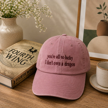 A dusty pink baseball cap with embroidered text reading 'you're all so lucky I don't own a dragon' sits on a wooden table next to a book, a cup, and decorative vases in a styled indoor setting.